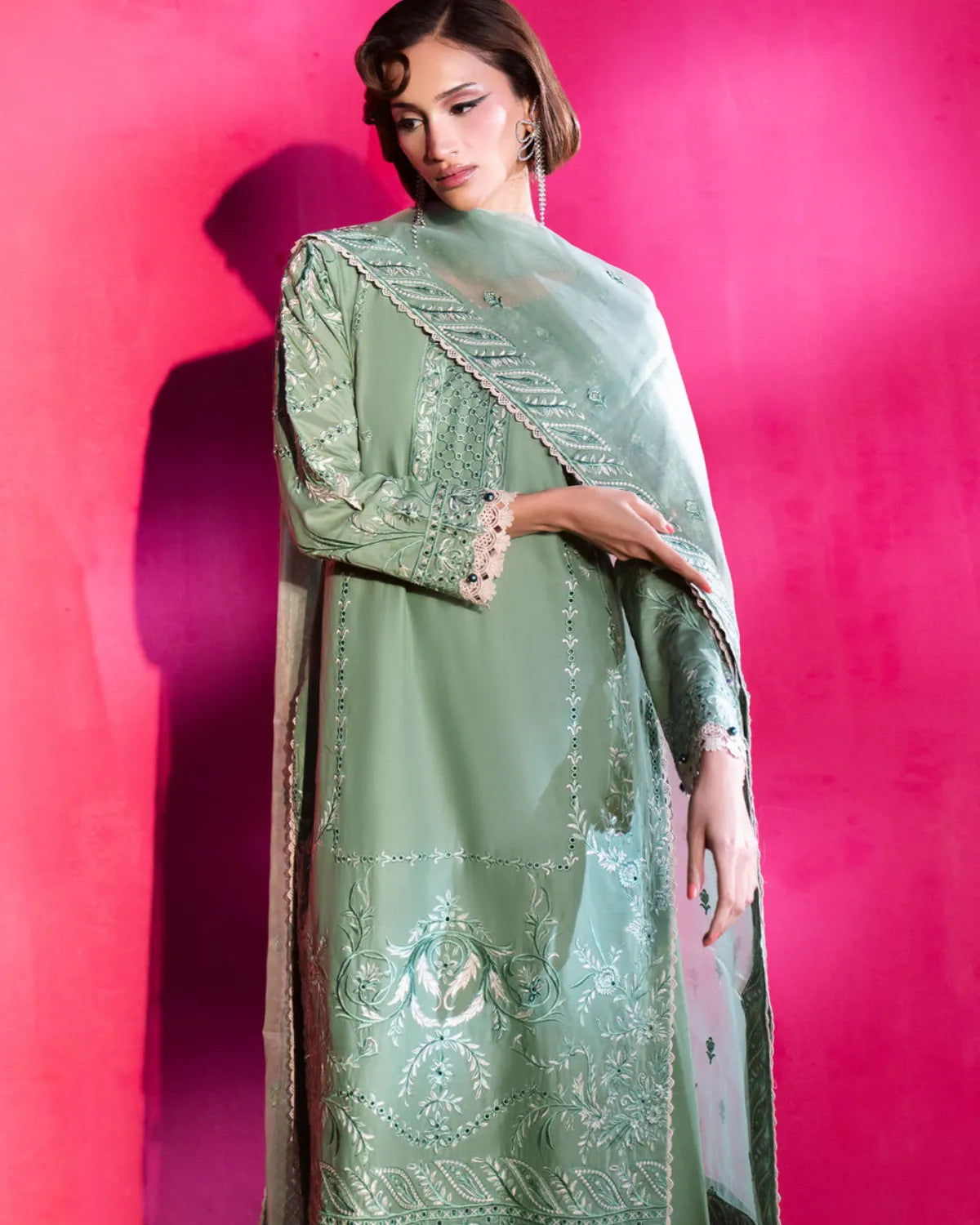 A woman models a sage green traditional outfit with intricate silver embroidery and a matching sheer dupatta against a vibrant pink background.