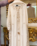 An ivory/cream long-sleeve abaya dress with intricate gold and beige floral embroidery, hanging in front of an ornate gold mirror.