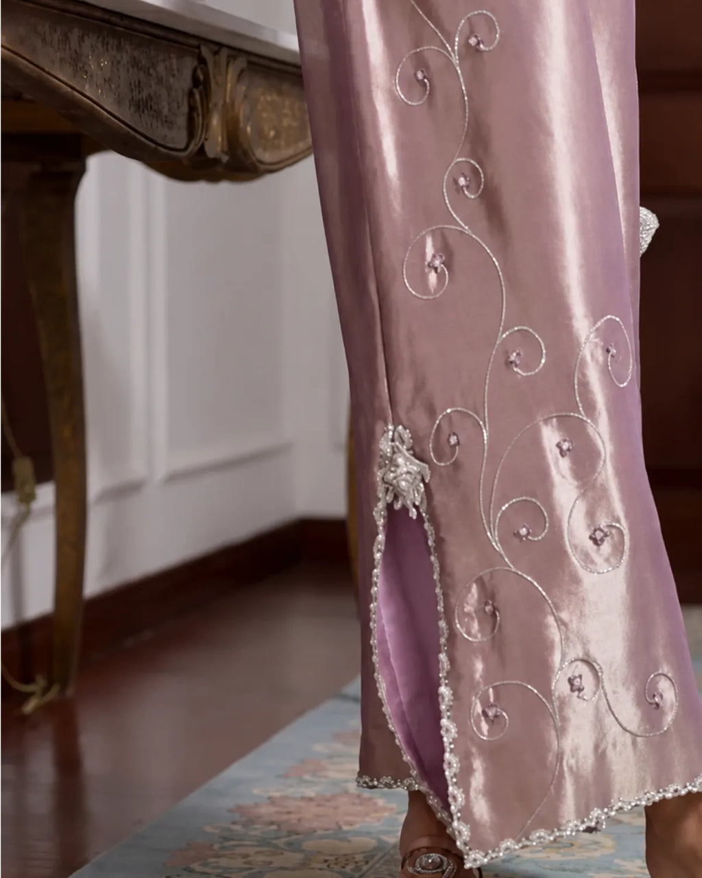 A close-up of a person wearing a lavender satin garment with silver embroidered swirl patterns and a silver beaded trim along the hem, standing in an indoor setting.