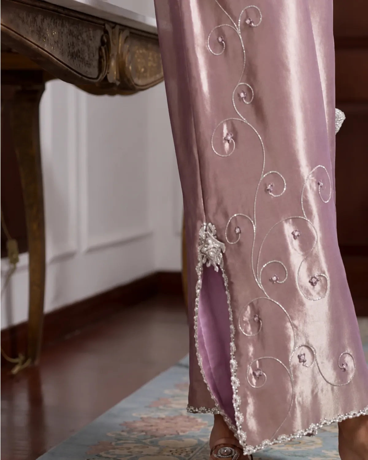 A close-up of a person wearing a lavender satin garment with silver embroidered swirl patterns and a silver beaded trim along the hem, standing in an indoor setting.