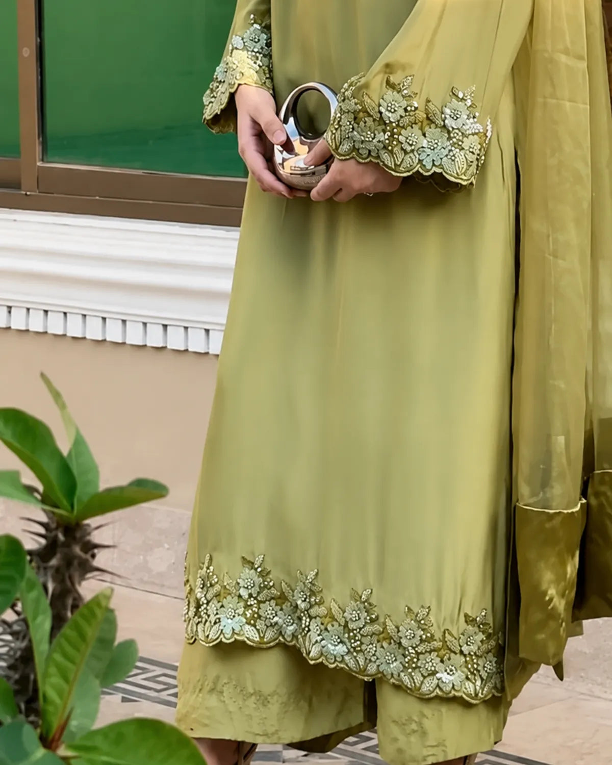 A person wearing a sage green traditional outfit with ornate floral embroidery on the cuffs and hem, holding a small metallic mirror. The background shows a beige wall, a green window frame, and some green plants.