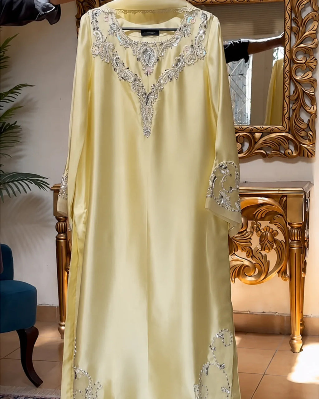 A pale yellow satin abaya dress with intricate silver and white embroidery around the V-neckline and sleeve cuffs, displayed in a boutique setting.