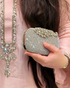 A close-up of a person holding a small, sparkling silver rhinestone clutch purse. The person is wearing a light pink embroidered traditional outfit with a matching necklace and bracelet.