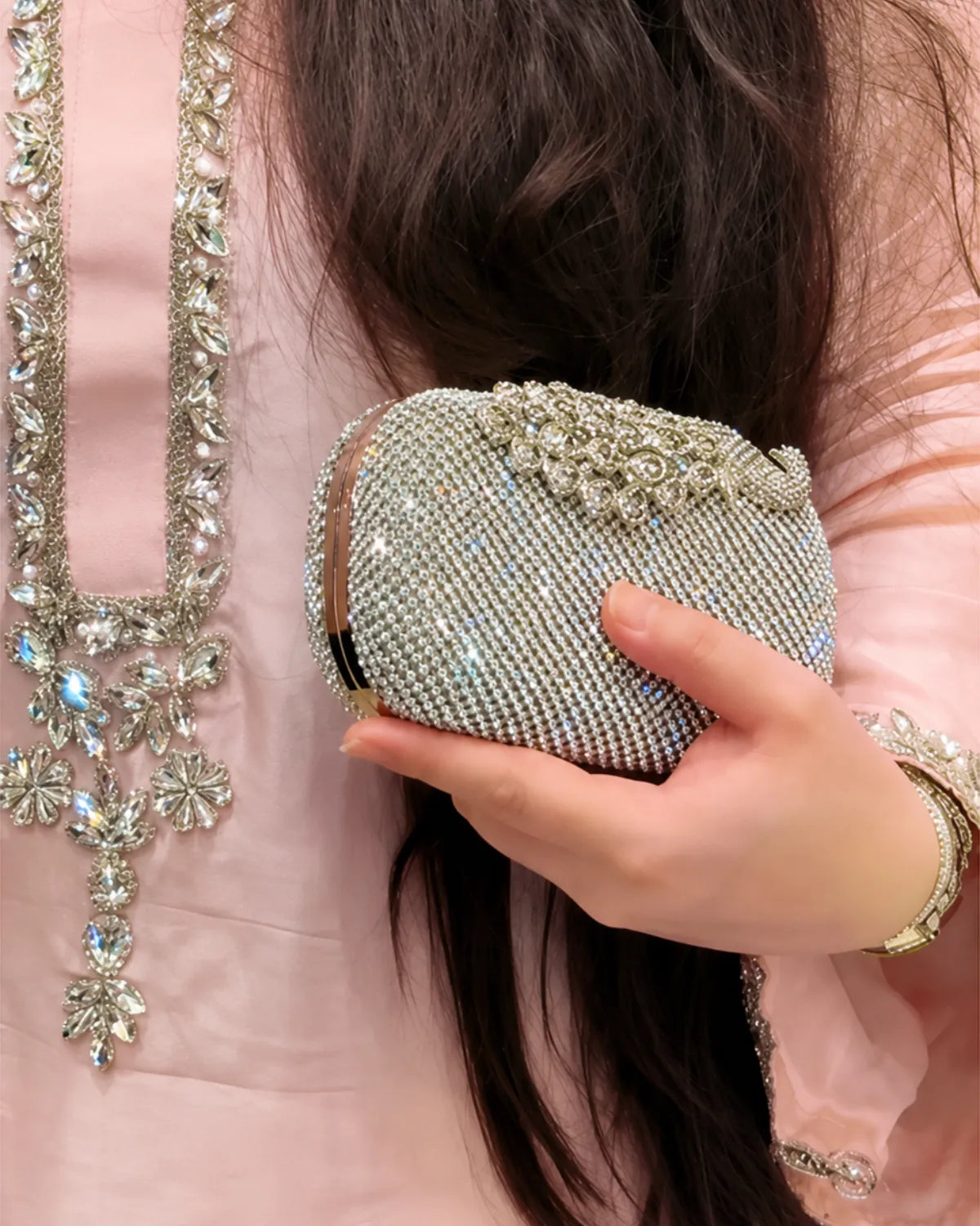 A close-up of a person holding a small, sparkling silver rhinestone clutch purse. The person is wearing a light pink embroidered traditional outfit with a matching necklace and bracelet.