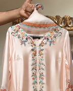 A pale blush pink satin long-sleeve top with colorful floral embroidery around the V-neck and down the center front, hanging on a hanger with a hand holding the collar.