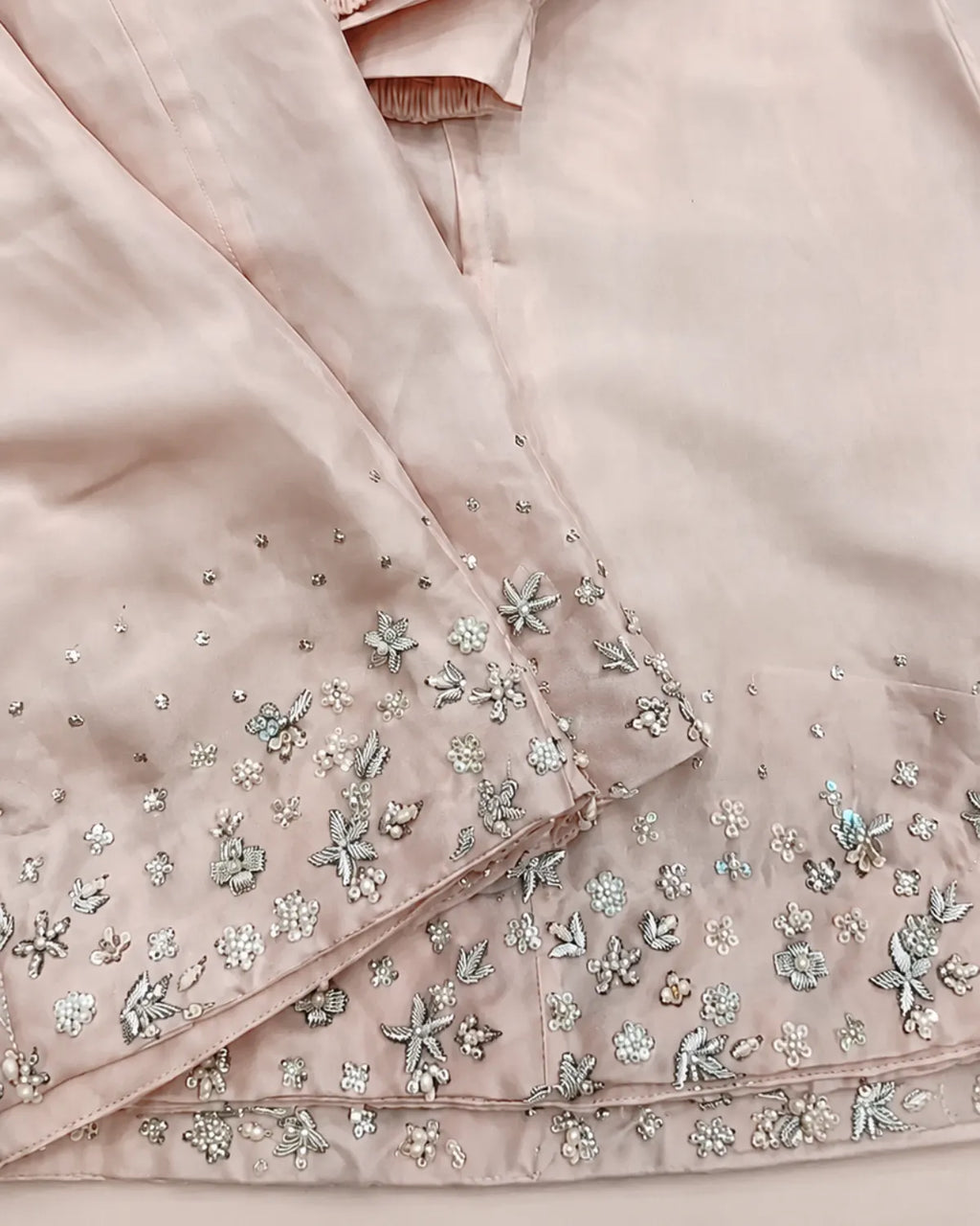 Close-up of a light blush pink fabric garment with detailed silver and white floral embroidery along the hem and sleeve.