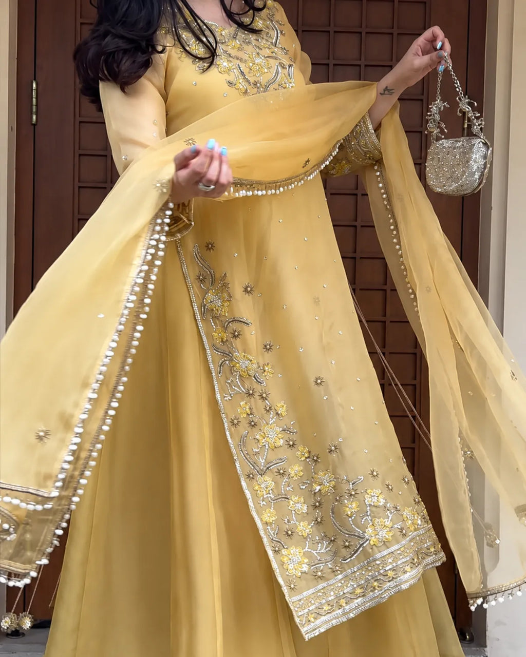 A woman wearing a traditional yellow anarkali gown with intricate gold and silver embroidery, holding a matching sheer dupatta and a small silver handbag.