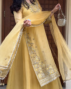 A woman wearing a traditional yellow anarkali gown with intricate gold and silver embroidery, holding a matching sheer dupatta and a small silver handbag.