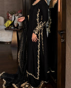 A black long gown with ornate gold floral embroidery and a matching sheer black dupatta, shown on a model in an indoor setting with a mirror reflecting the back of the dress.