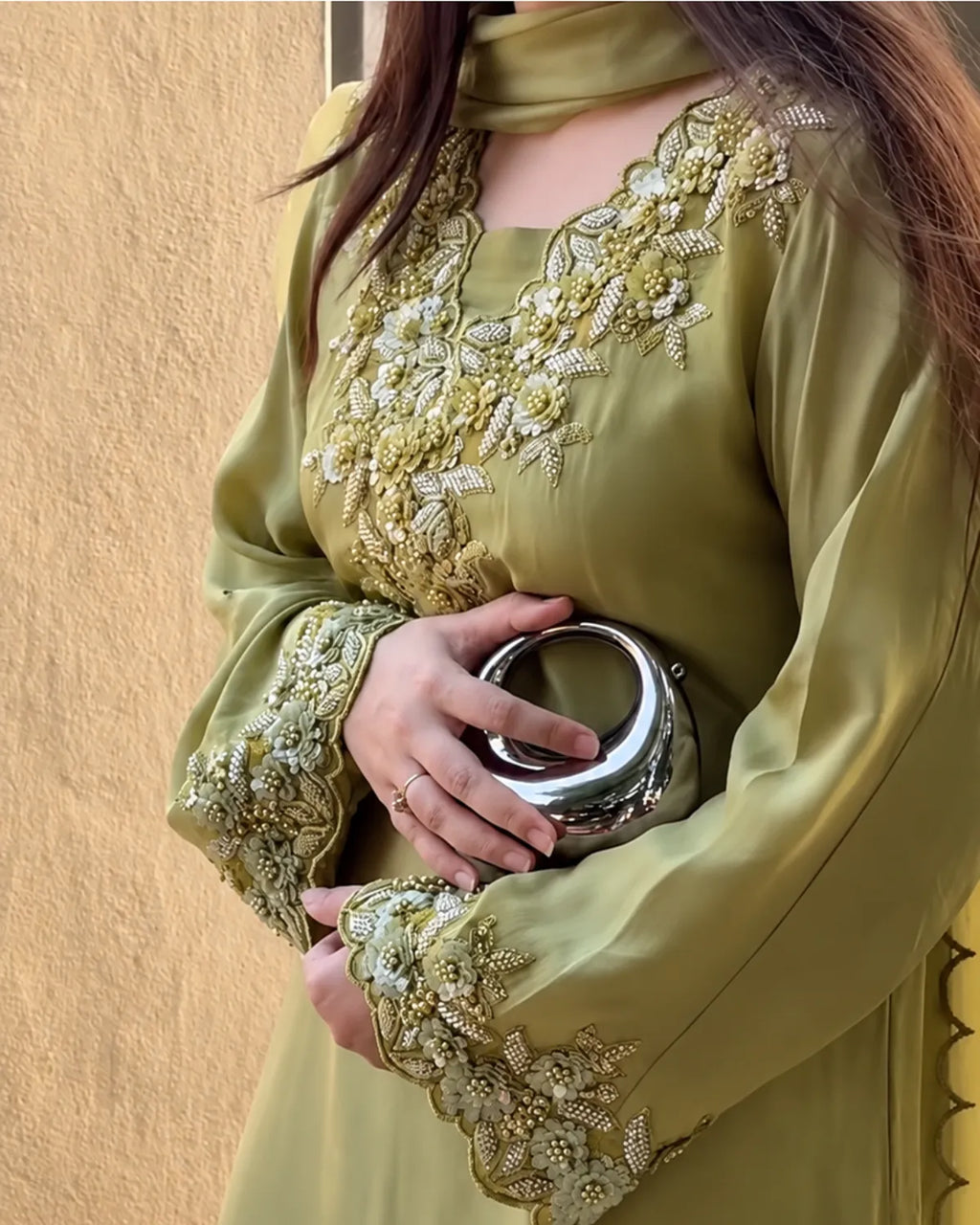 A woman wearing a sage green traditional long-sleeve dress with intricate gold and white floral embroidery on the neckline and cuffs, holding a metallic silver clutch bag against a textured beige wall.