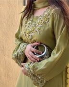 A woman wearing a sage green traditional long-sleeve dress with intricate gold and white floral embroidery on the neckline and cuffs, holding a metallic silver clutch bag against a textured beige wall.