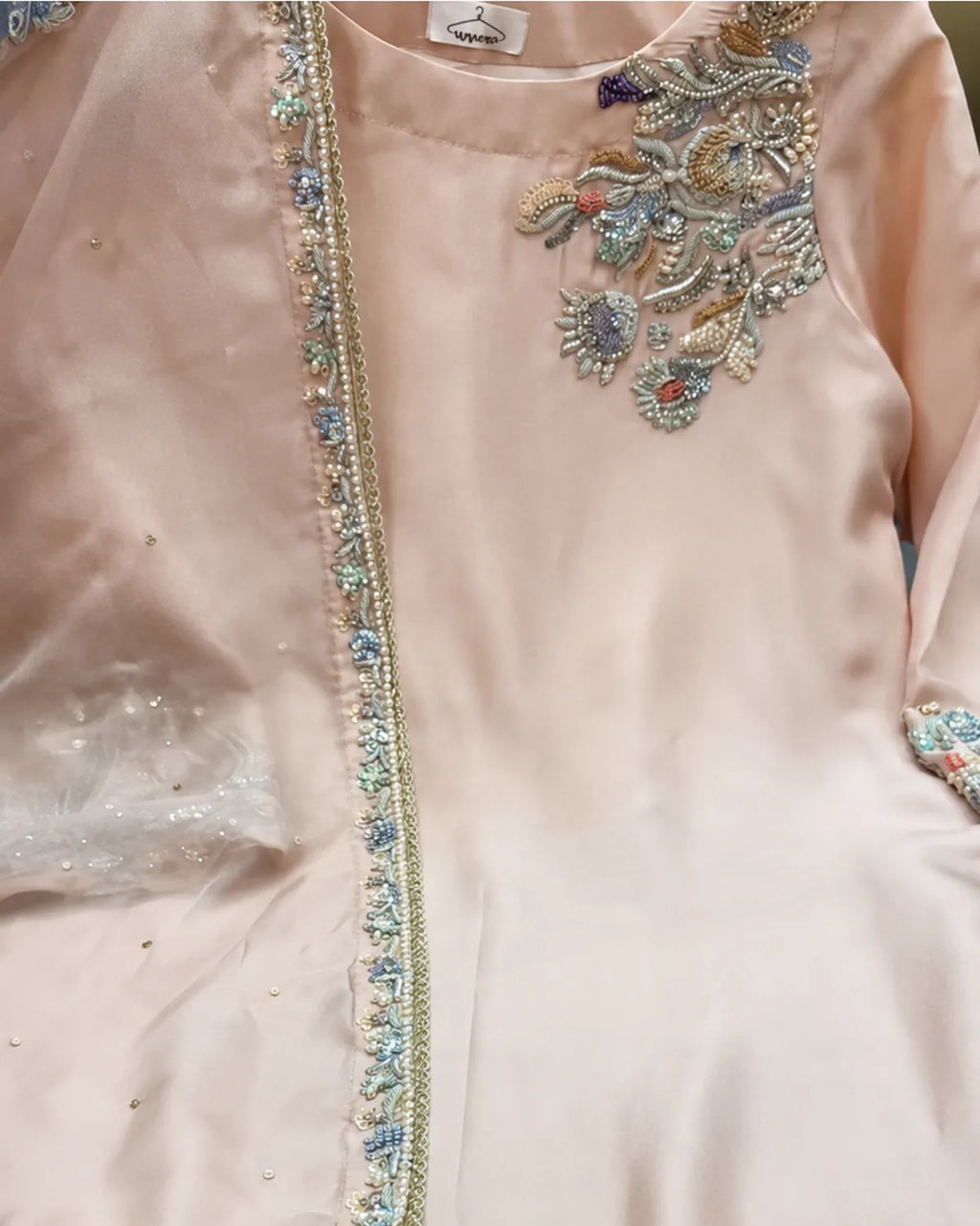 A close-up of a light peach-pink traditional embroidered garment, likely a kurta or salwar suit piece, featuring ornate metallic and beadwork embroidery on the chest and a decorative border along the front opening.