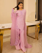 A woman modeling a light pink traditional South Asian outfit with matching wide-leg pants and a sheer dupatta, standing in an indoor setting with marble-topped tables.