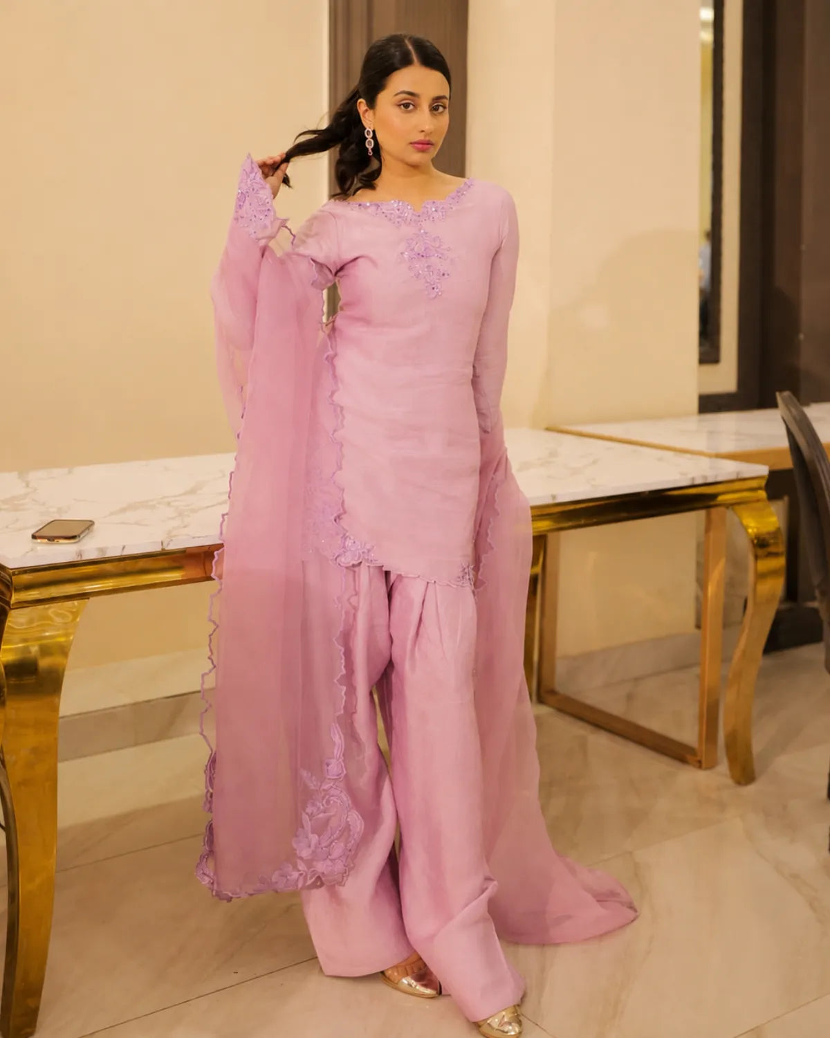 A woman modeling a light pink traditional South Asian outfit with matching wide-leg pants and a sheer dupatta, standing in an indoor setting with marble-topped tables.