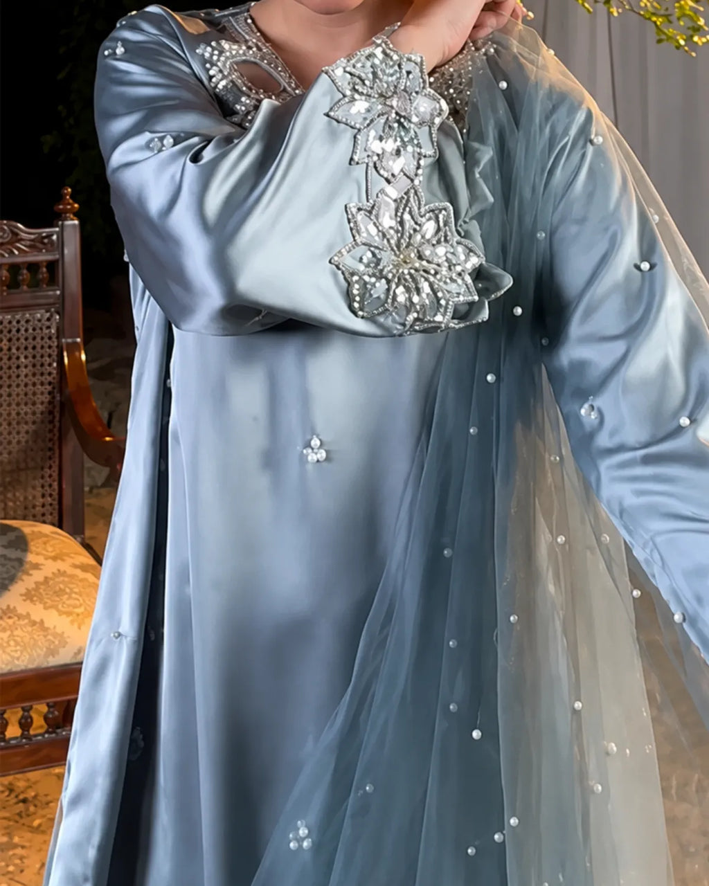 A person wearing a light blue satin traditional dress with ornate silver embellishments on the chest and shoulders, paired with a sheer matching dupatta decorated with small pearl-like beads.