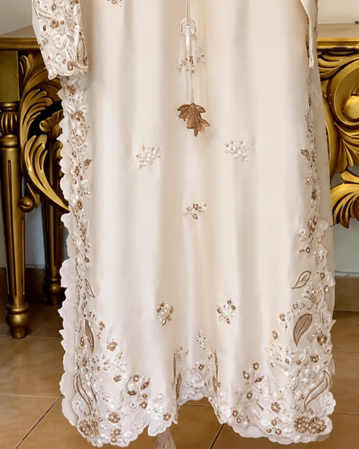 An ivory or cream-colored dress with intricate floral embroidery and lace trim, featuring a central hanging tassel ornament, shown hanging in front of a gold-colored ornate furniture piece.
