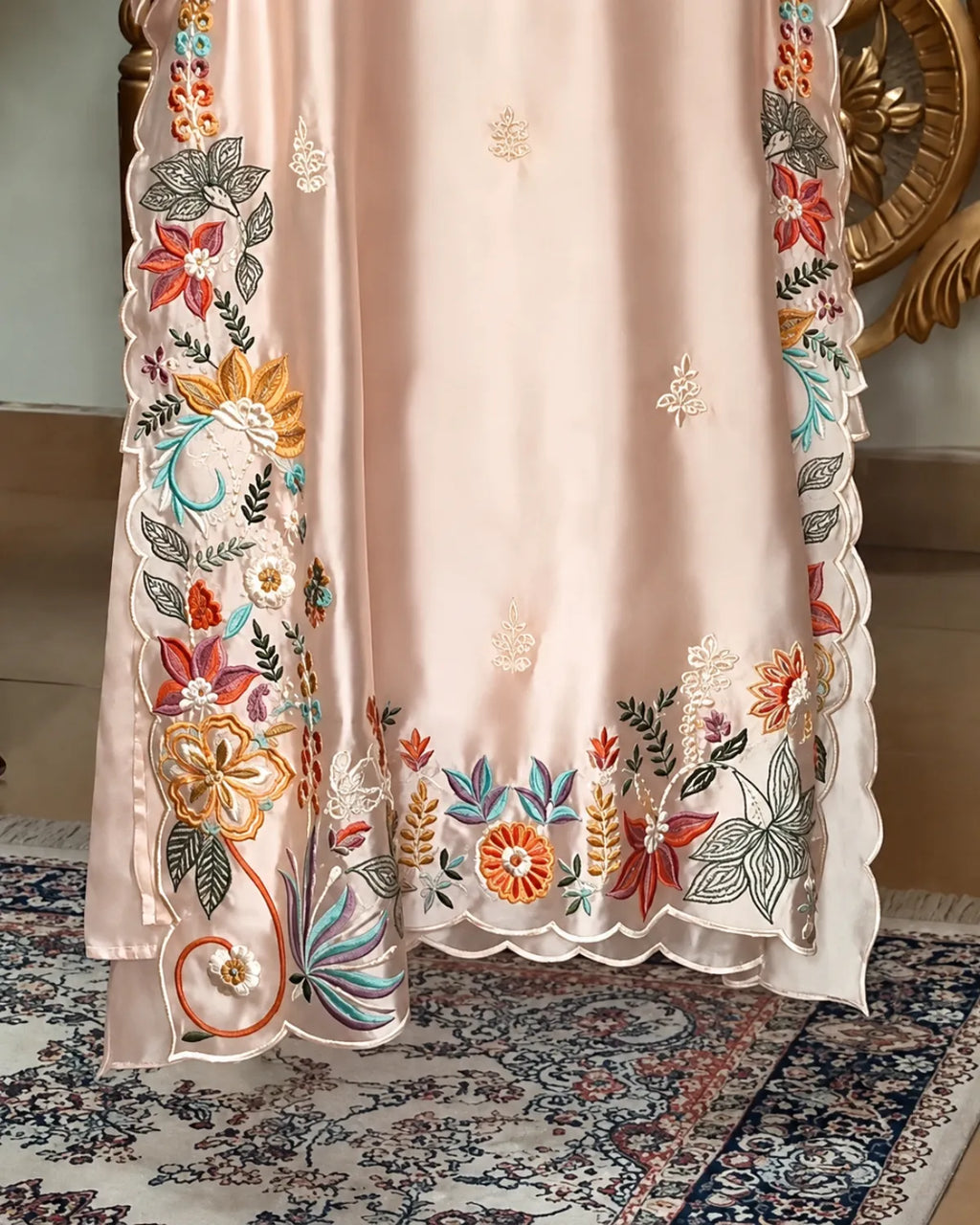 A close-up of a light blush pink embroidered fabric, likely a traditional dress or dupatta, featuring colorful floral and leaf embroidery along the borders and scattered motifs on the center. The fabric is draped over a chair, with a patterned rug and a gold-toned chair back visible in the background.