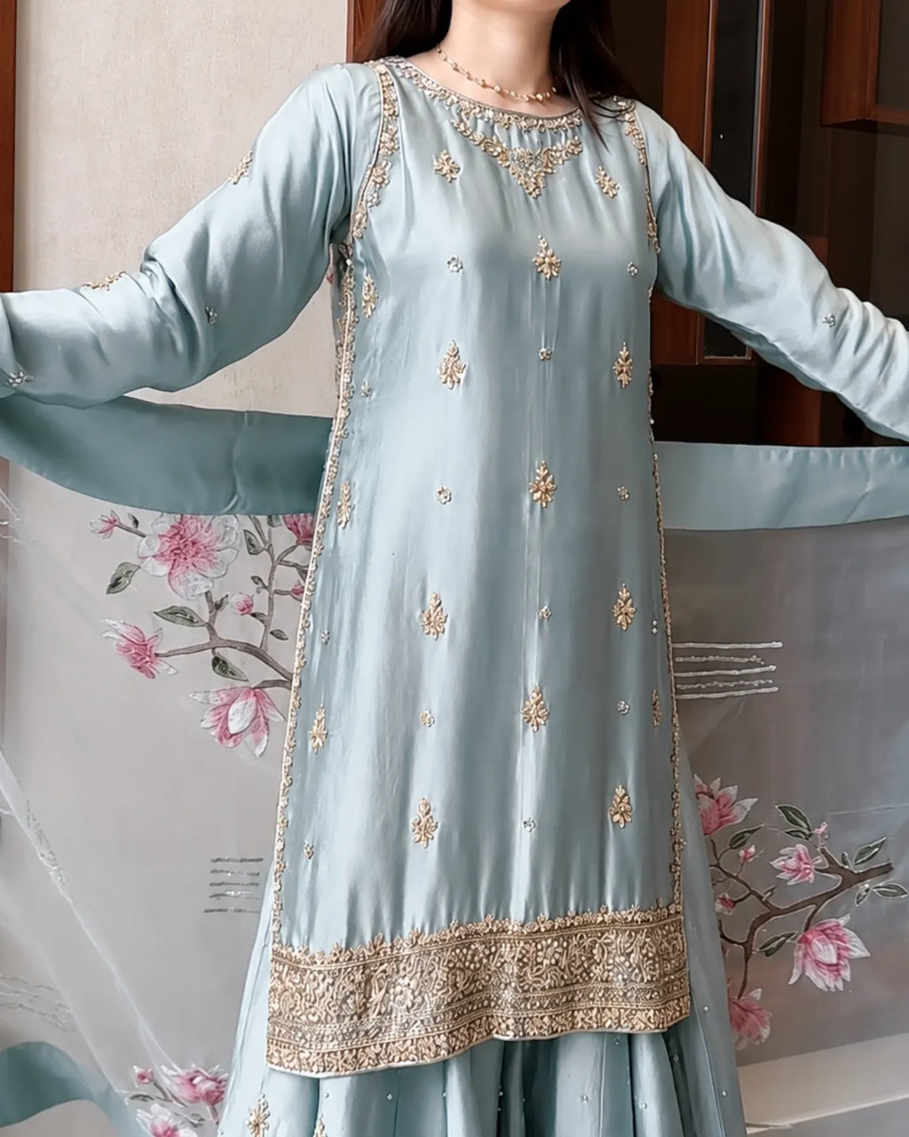 A woman wearing a light blue-green salwar kameez suit with gold embroidery and a matching sheer dupatta with a pink floral print.