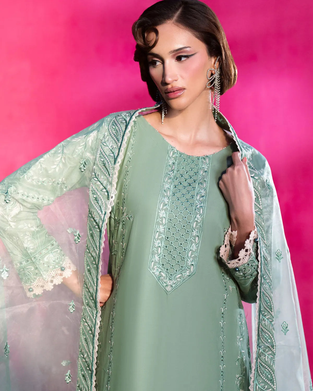 A woman modeling a sage green embroidered kurta with a matching sheer white dupatta featuring green embroidery, set against a vibrant pink studio background.