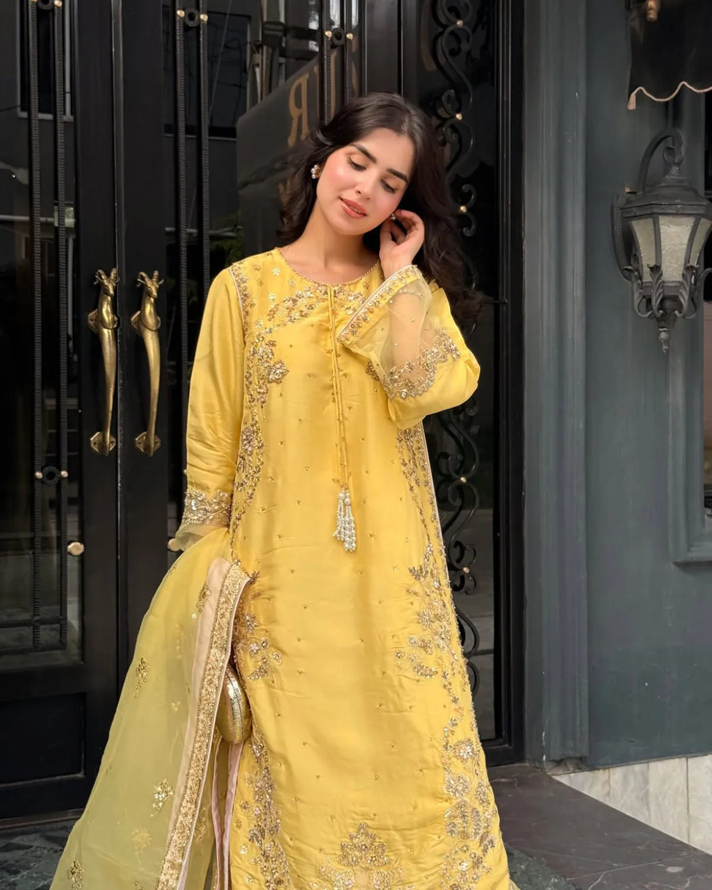 A woman wearing a bright yellow traditional embroidered outfit with a matching dupatta, standing in front of a dark ornate doorway.