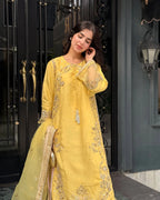 A woman wearing a bright yellow traditional embroidered outfit with a matching dupatta, standing in front of a dark ornate doorway.