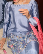 A woman wearing a light blue satin kurta with floral embroidery on the skirt and a bright pink dupatta draped over her shoulder.