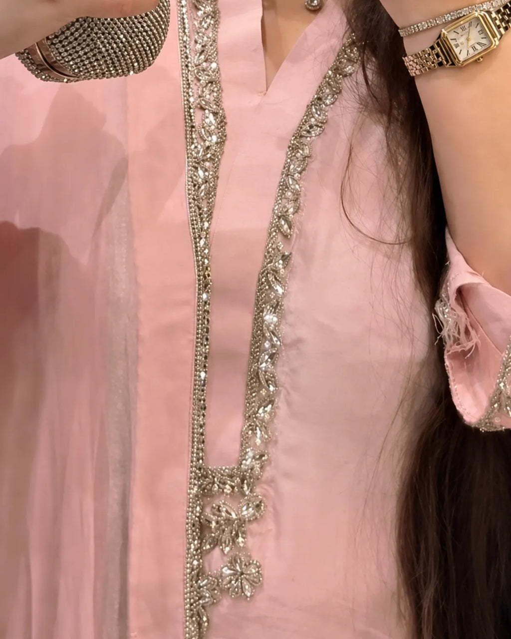 A close-up of a light pink traditional outfit with ornate silver and crystal embellishments along the neckline and front opening. A person is wearing a rhinestone-embellished clutch and a gold-tone watch, with long dark hair visible.