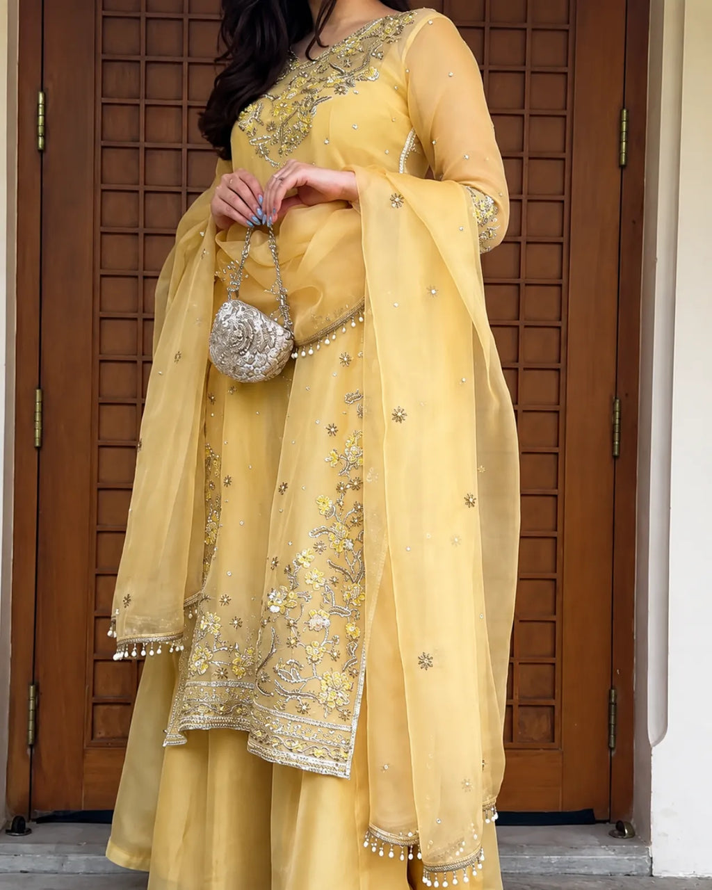 A woman wearing a traditional Indian outfit in light yellow with intricate silver and gold embroidery, holding a small silver purse in front of a brown wooden door.