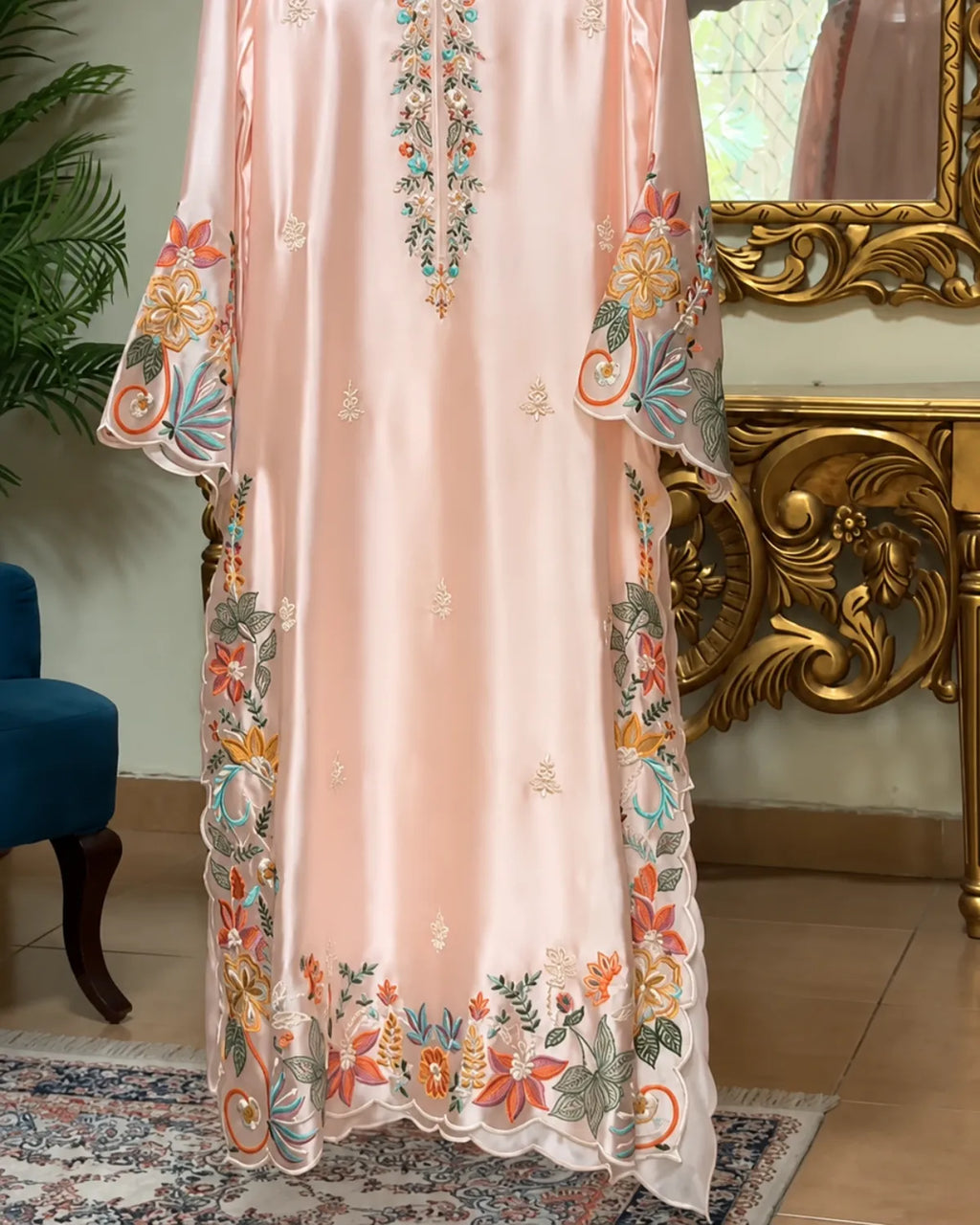 A light peach-pink satin or silk kaftan dress with colorful floral embroidery and scalloped lace trim, shown hanging in an ornate room with a gold console table and mirror.