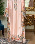 A light peach-pink satin or silk kaftan dress with colorful floral embroidery and scalloped lace trim, shown hanging in an ornate room with a gold console table and mirror.
