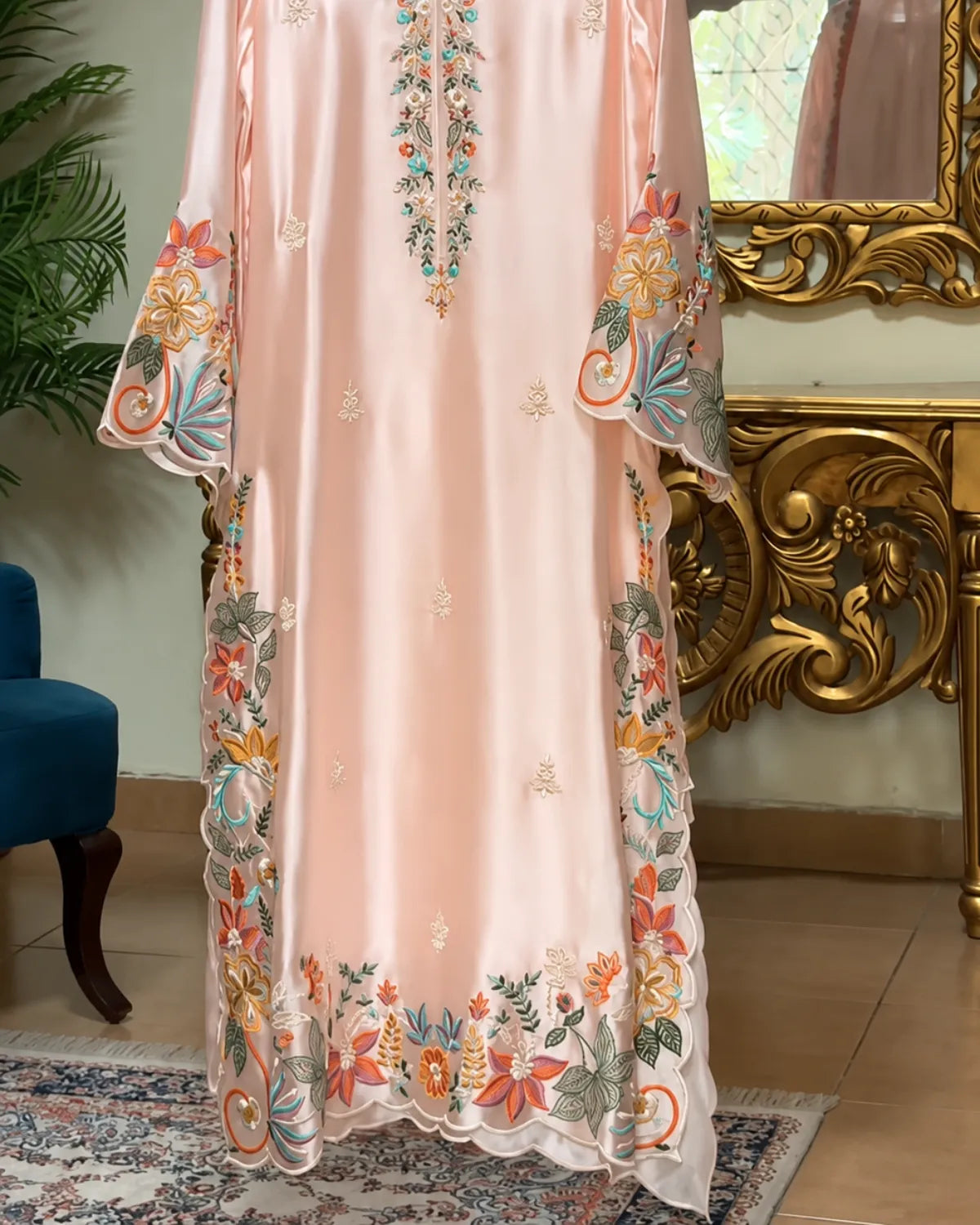 A light peach-pink satin or silk kaftan dress with colorful floral embroidery and scalloped lace trim, shown hanging in an ornate room with a gold console table and mirror.