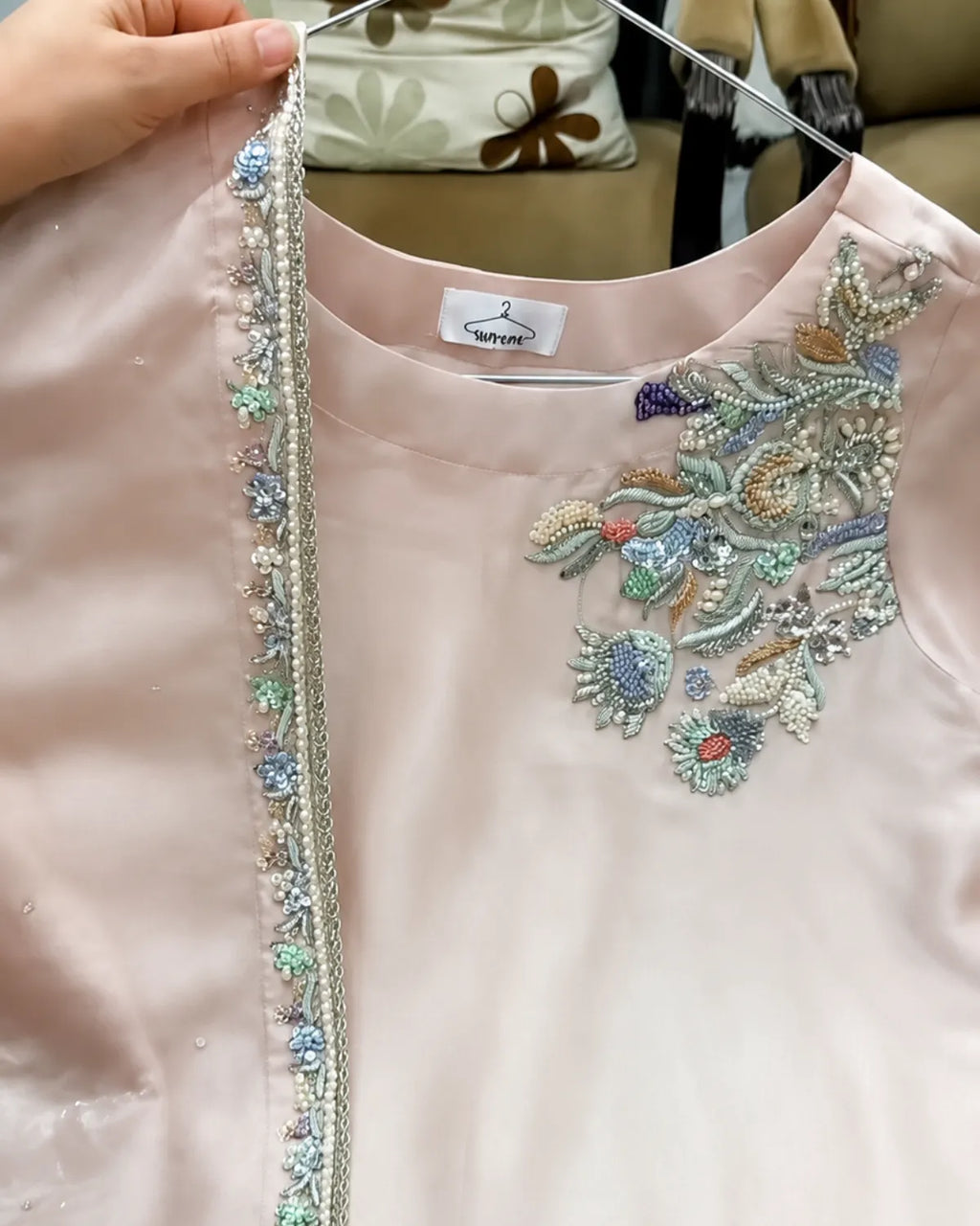A close-up of a light pink embroidered garment held up by a hand on a hanger, showing detailed multicolor beadwork and sequin embellishments on the chest and neckline.