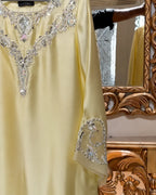 A pale yellow satin abaya dress with intricate silver crystal embroidery on the V-neckline and sleeve, shown hanging in front of an ornate gold mirror.