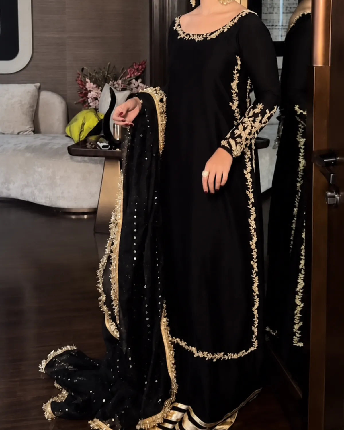 A woman wearing a black traditional Anarkali gown with ornate gold embroidery and a matching black dupatta with gold trim, standing in a furnished indoor room.