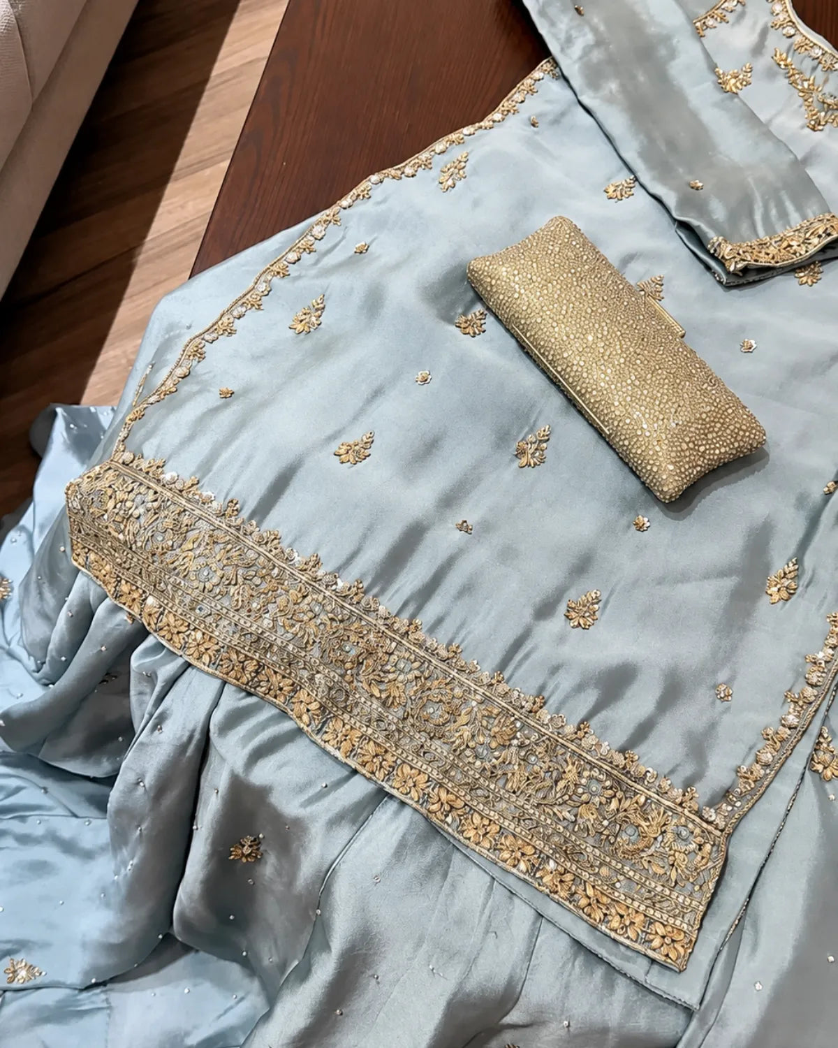 A light blue satin ethnic outfit with gold embroidery and a matching gold clutch purse, laid out on a dark wooden surface.