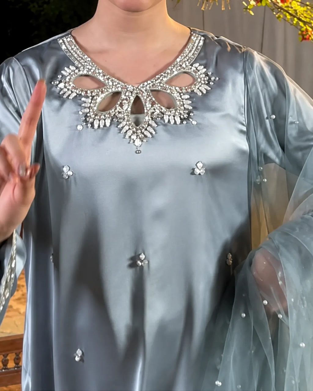A close-up of a person wearing a light blue satin tunic top with a heavily embellished silver rhinestone V-neckline and small scattered rhinestone accents on the fabric and sheer sleeves.