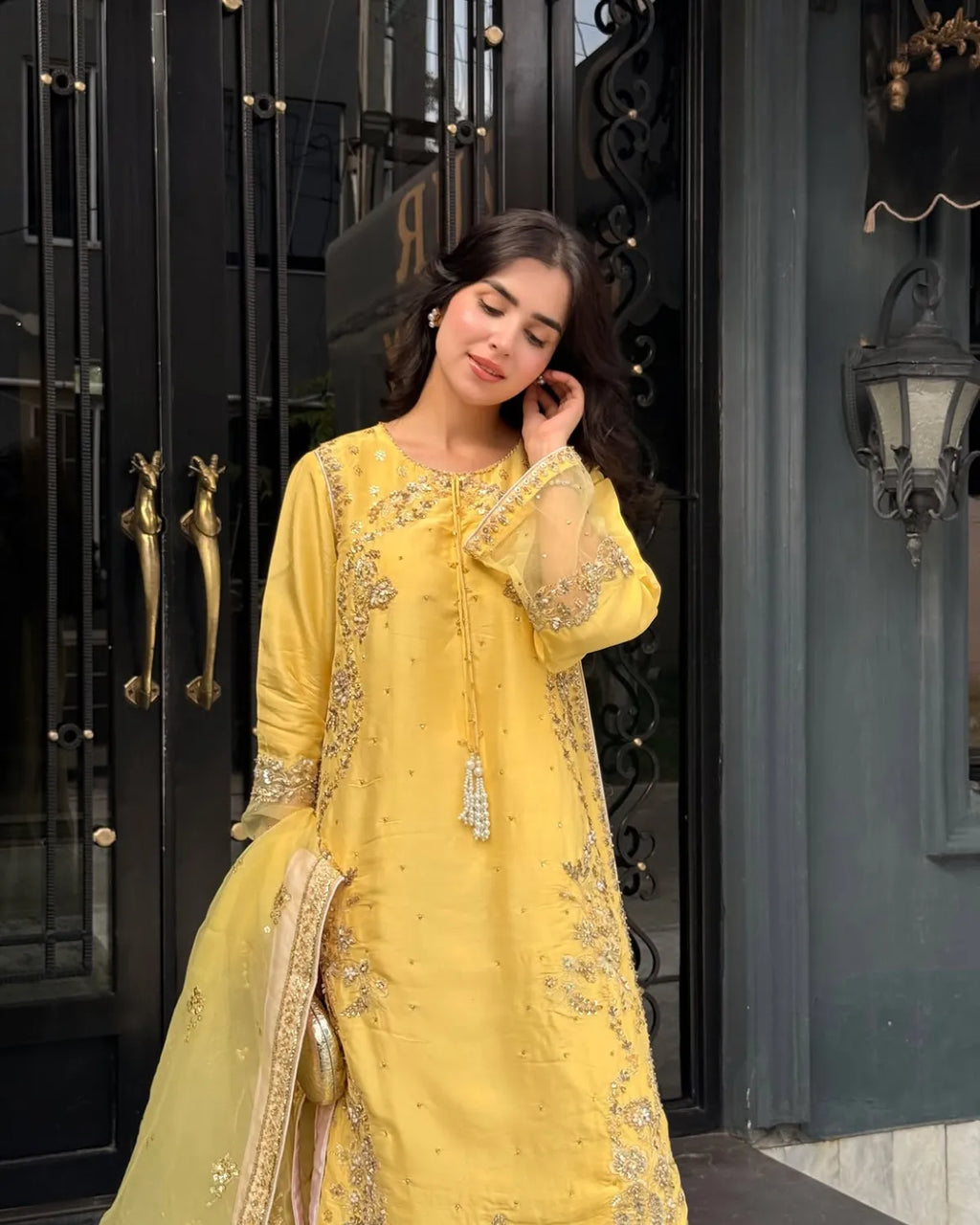 A woman wearing a bright yellow embroidered traditional outfit with a matching dupatta, standing in front of a black metal gate and dark wall.