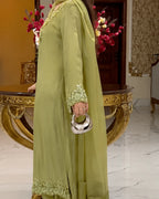 A woman wearing a light olive green traditional outfit with floral embroidery on the neckline, cuffs, and hem, holding a small metallic clutch in an ornate indoor setting.