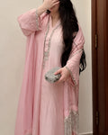 A woman wearing a light pink traditional outfit with a matching long sheer outer layer decorated with silver beaded trim and fringe, holding a silver rhinestone clutch against an indoor background.