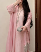 A woman wearing a light pink traditional outfit with a matching long sheer outer layer decorated with silver beaded trim and fringe, holding a silver rhinestone clutch against an indoor background.