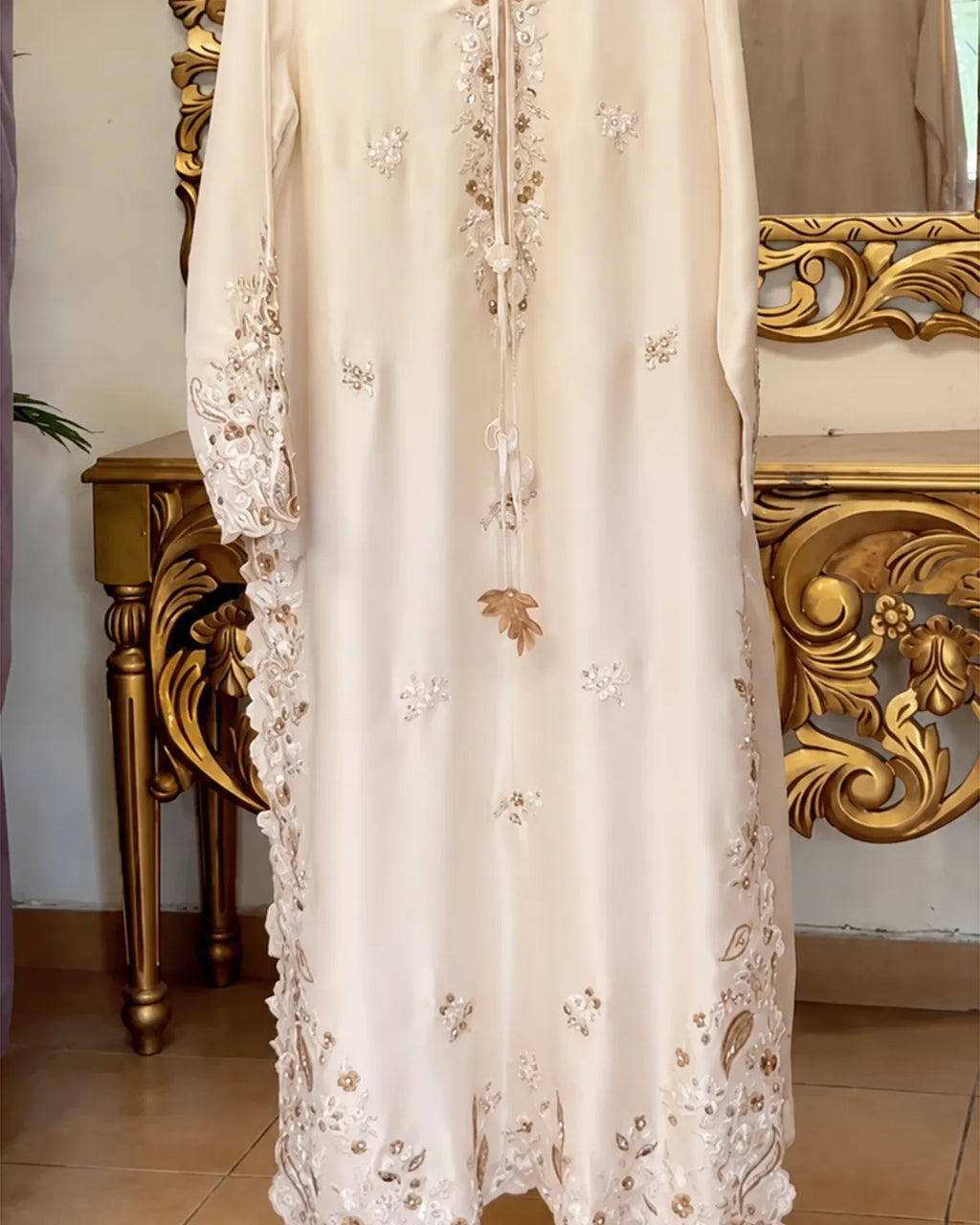 An ivory or cream-colored long dress with intricate floral embroidery and lace detailing, shown hanging in front of an ornate gold mirror.
