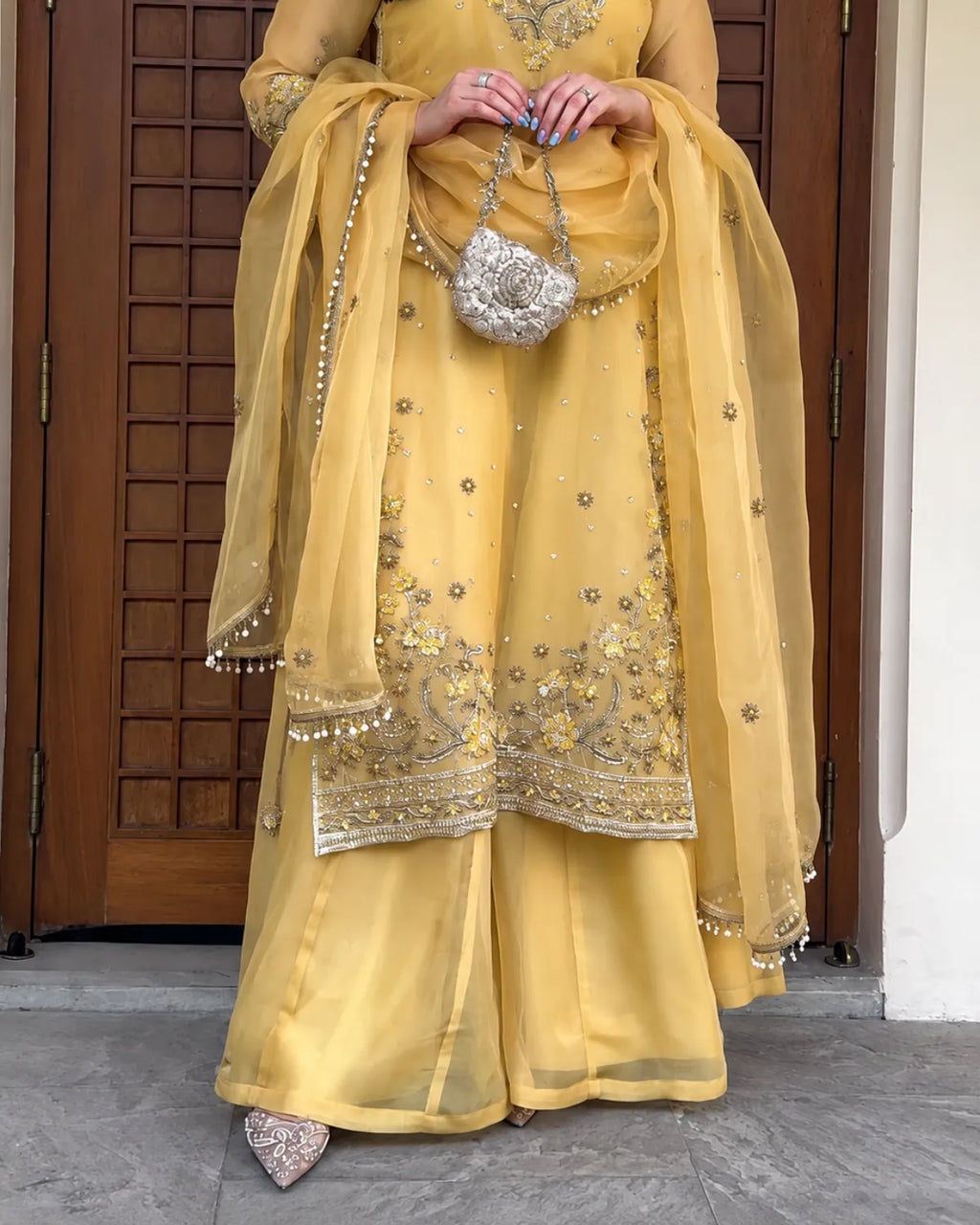 A person wearing a yellow traditional outfit with silver embroidery and a matching sheer dupatta, holding a small silver handbag, standing in front of a brown wooden door.