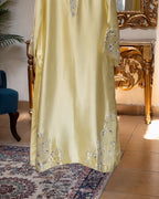 A pale yellow satin or silk kaftan dress with intricate silver and white beadwork embroidery, shown hanging in an elegant room with a gold console table and patterned rug.