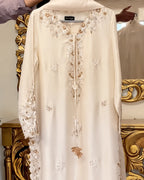 A cream-colored long-sleeve tunic with intricate gold and white floral embroidery, hanging in front of an ornate gold mirror.