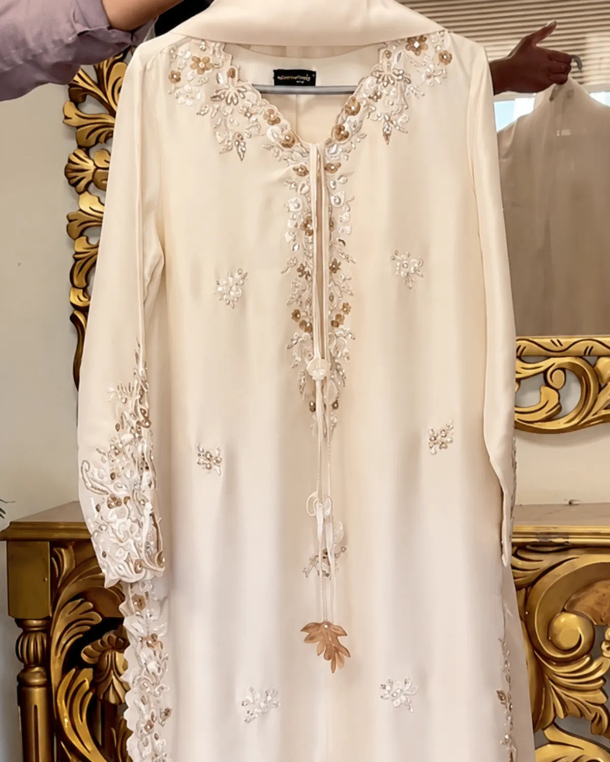 A cream-colored long-sleeve tunic with intricate gold and white floral embroidery, hanging in front of an ornate gold mirror.