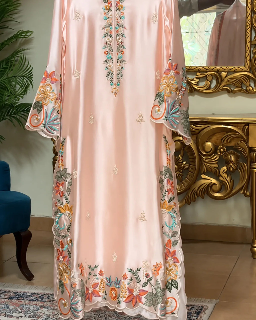 A long peach-pink satin caftan with colorful floral embroidery, displayed in an ornate room with a gold console table and mirror.
