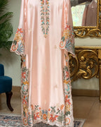A long peach-pink satin caftan with colorful floral embroidery, displayed in an ornate room with a gold console table and mirror.