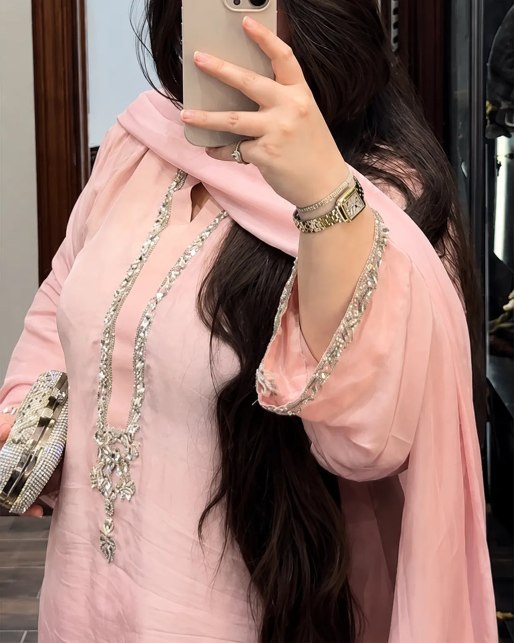 A woman takes a mirror selfie wearing a light pink traditional outfit with silver embellished trim. She is holding a silver patterned clutch purse and wearing a silver watch.
