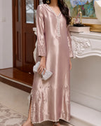 A woman wearing a long, shiny blush pink kaftan dress with silver embroidery and beadwork along the neckline and hem, holding a small silver clutch in an indoor setting.