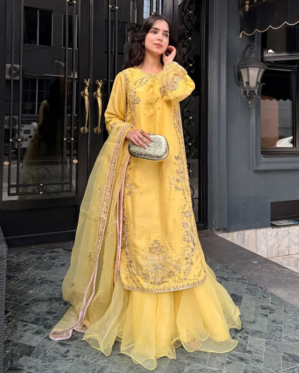 A woman models a traditional yellow Indian salwar kameez suit with intricate silver embroidery and a matching sheer dupatta, standing in front of a dark building entrance.